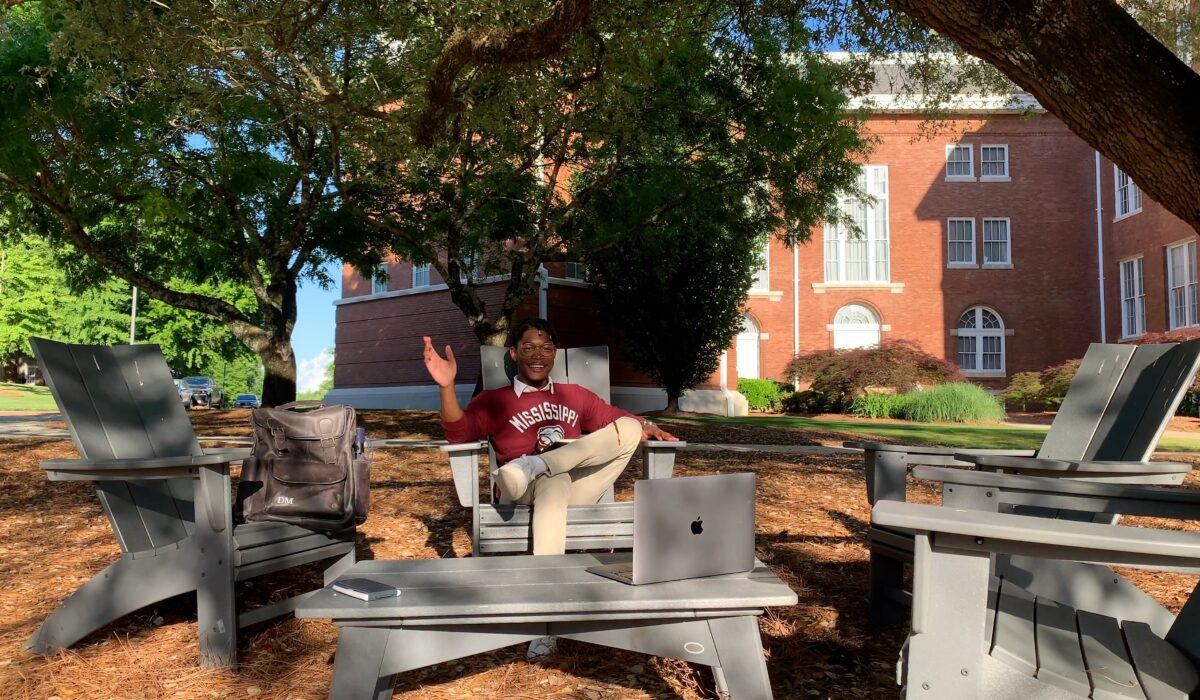 Image of Nico Miller smiling whilst sitting on chairs in front of a brick building.